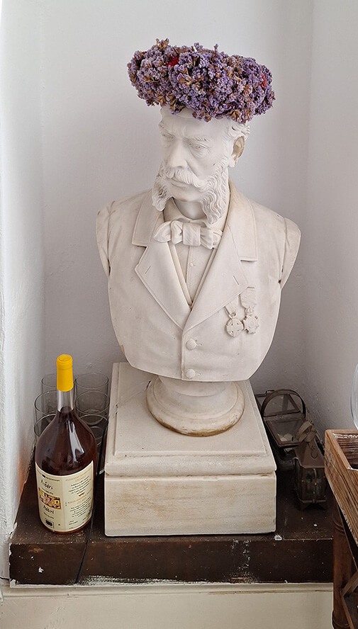 Προτομή του Γεωργίου Ε. Βέλτσου υπό Γεωργίου Βιτάλη. Αντίγραφο του έργου, όπως συνηθίζονταν, κατείχε η οικογένεια  από γενιά σε γενιά .Εδω, ο Γεώργιος Βελτσος, στεφανομένος με  μοβ αμάραντα,  συγκατοικεί  προ ογδονταετίας και πλέον με τον Γιώργο Βέλτσο στην οικία του στη Μύκονο ― δίχως να θέτει ζητήματα αυθεντικού και αντιγράφου.